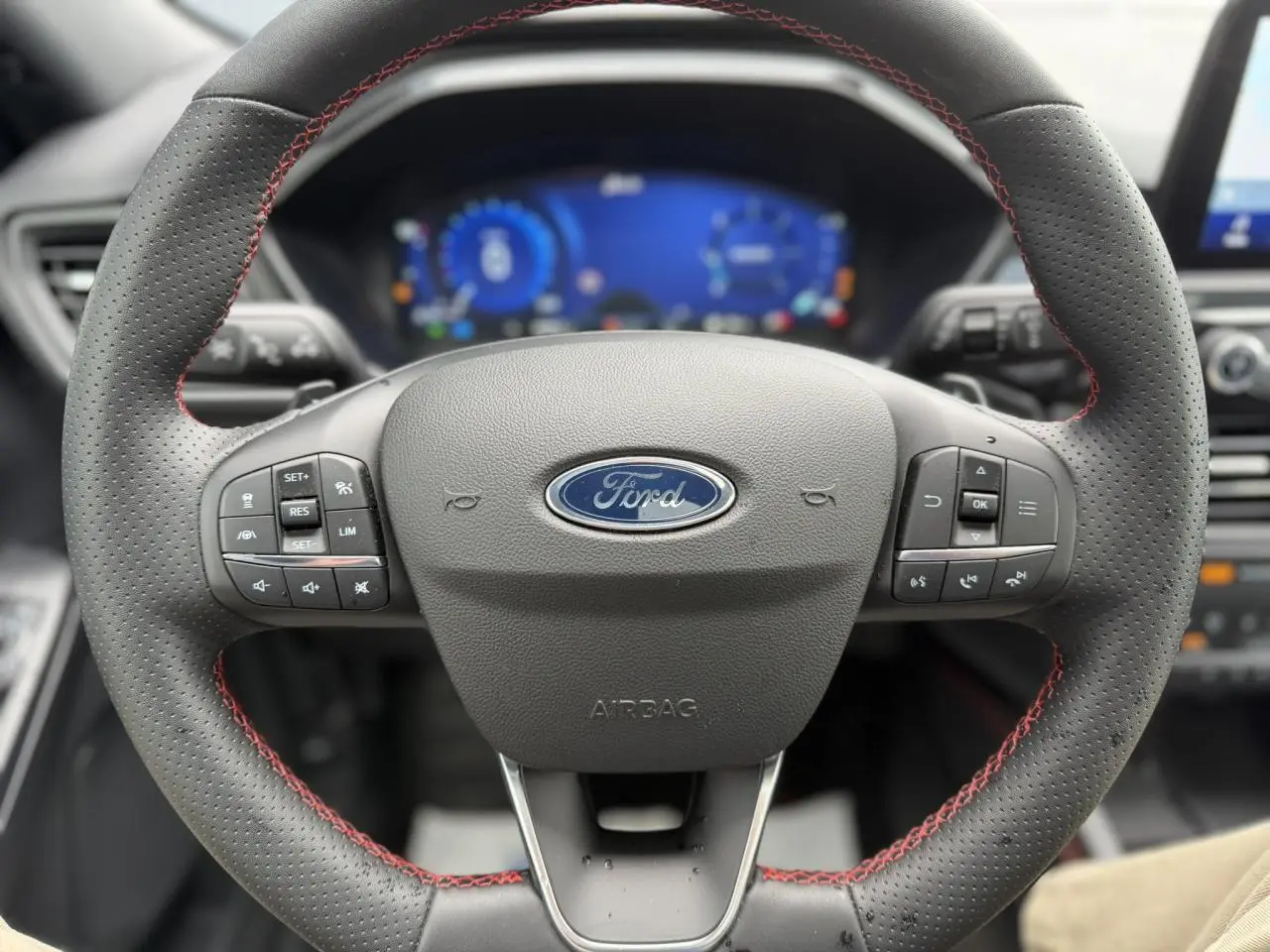 Vue rapprochée du volant perforé noir avec surpiqûres rouges du Ford Kuga 2023, tableau de bord numérique en arrière-plan.