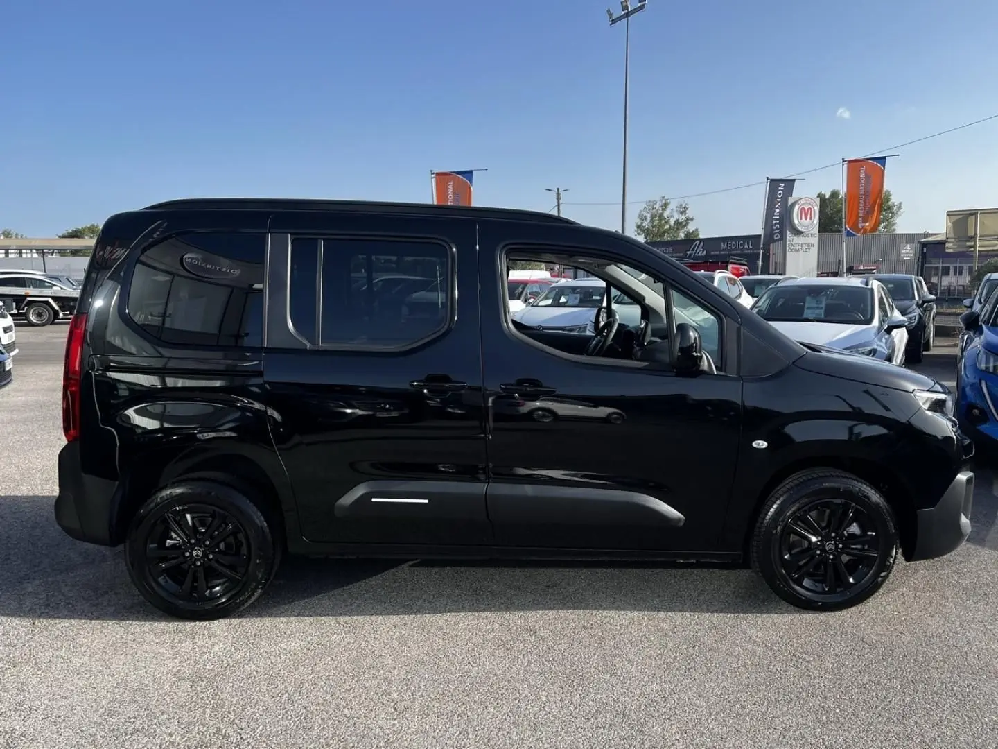 Vue latérale droite d'un Citroën Berlingo noir Perla Nera avec jantes noires et vitres teintées, sur parking extérieur.