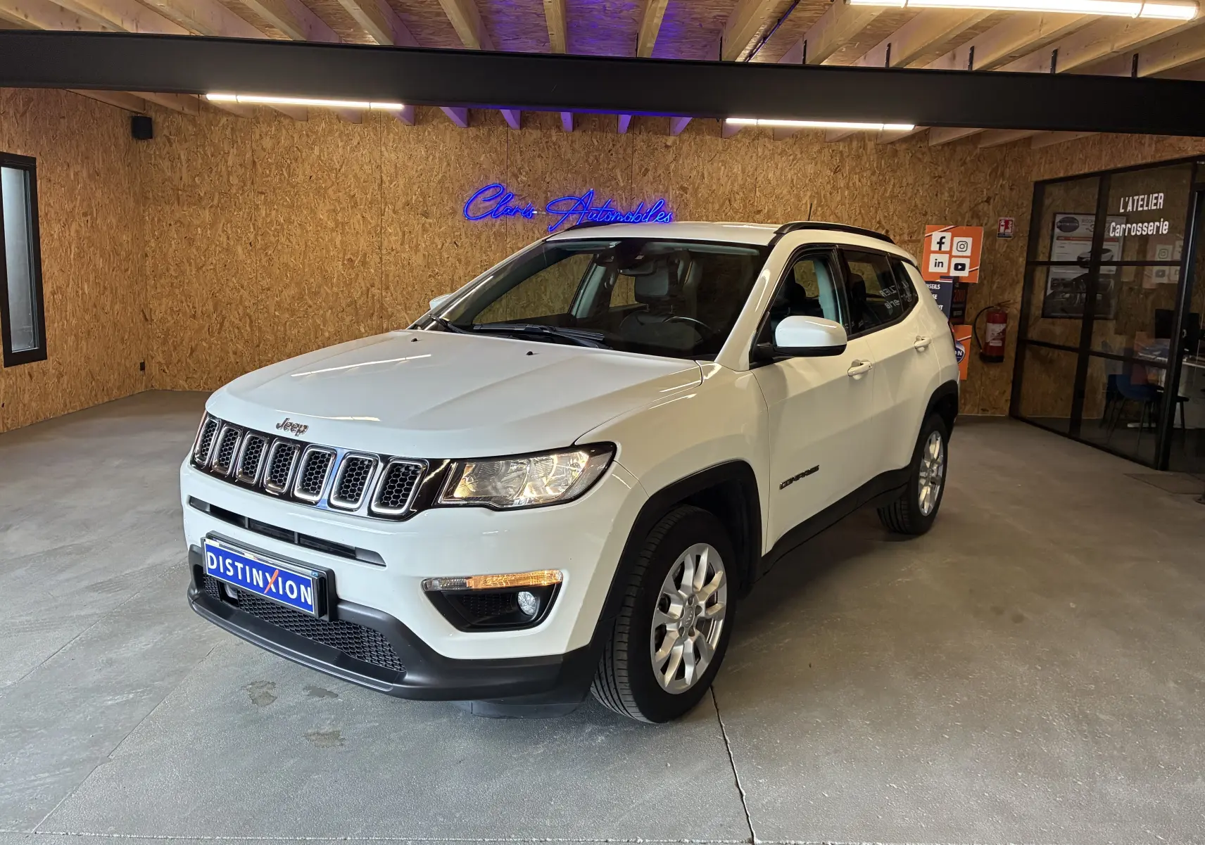 Jeep Compass ivoire en 3/4 avant droit, avec barres de toit et jantes alu, stationné en intérieur sur sol béton.