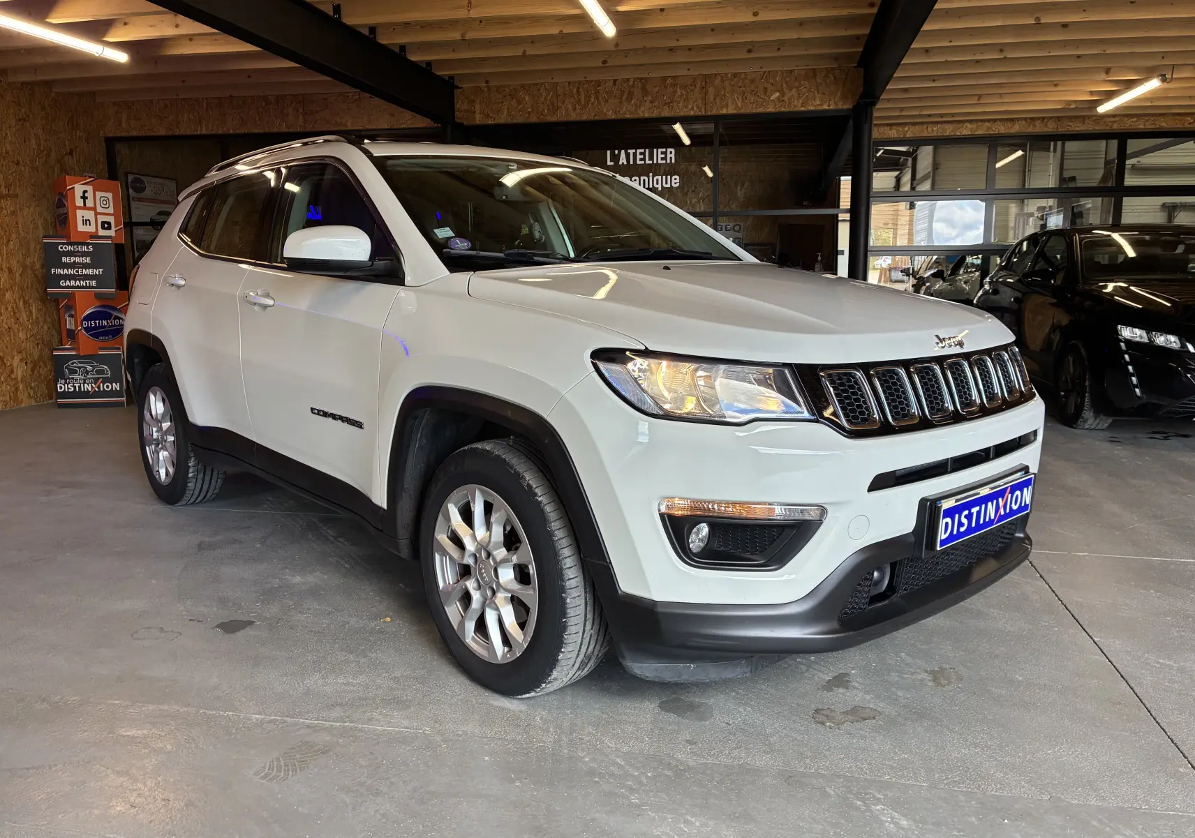 Jeep Compass ivoire en 3/4 avant droit, avec barres de toit et jantes alu visibles en intérieur d'atelier.