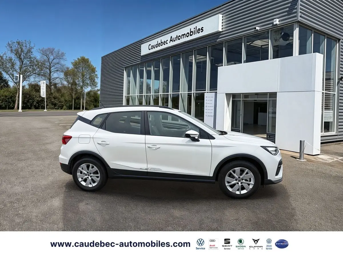 Vue de profil côté gauche d'un SEAT Arona blanc garé devant un bâtiment moderne de concession automobile.