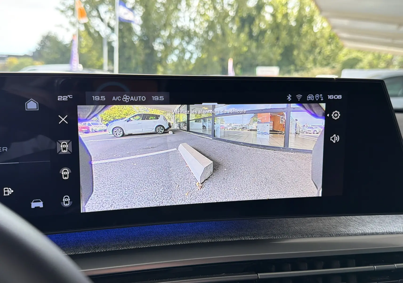 Écran intérieur montrant la caméra de recul d'une Peugeot 1007 gris artense avec vue sur un parking et un trottoir.