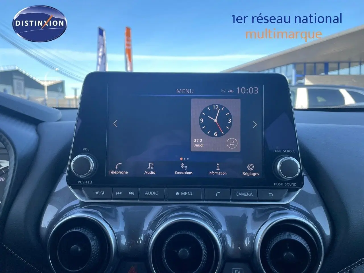 Écran tactile central du tableau de bord du Nissan Juke gris, affichant l'horloge et les options multimédia.