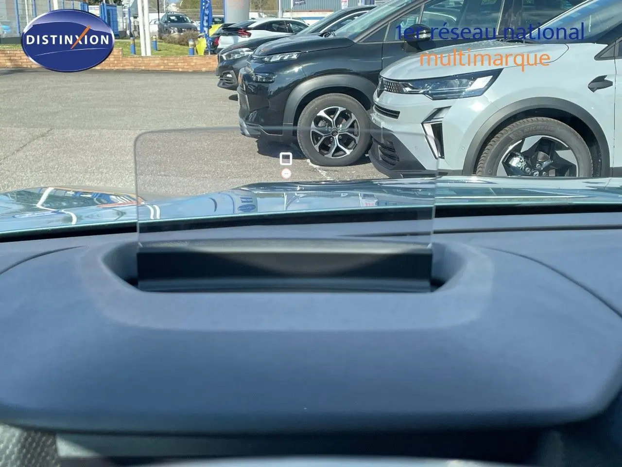Vue depuis l'intérieur sur le tableau de bord et le pare-brise, avec plusieurs SUV gris et noir en stationnement devant.