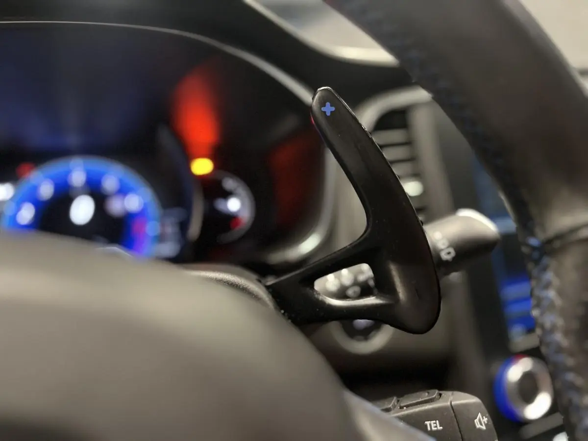 Gros plan sur la palette de changement de vitesse + au volant cuir perforé de la Renault Mégane GT bleue, tableau de bord flou en arrière-plan.
