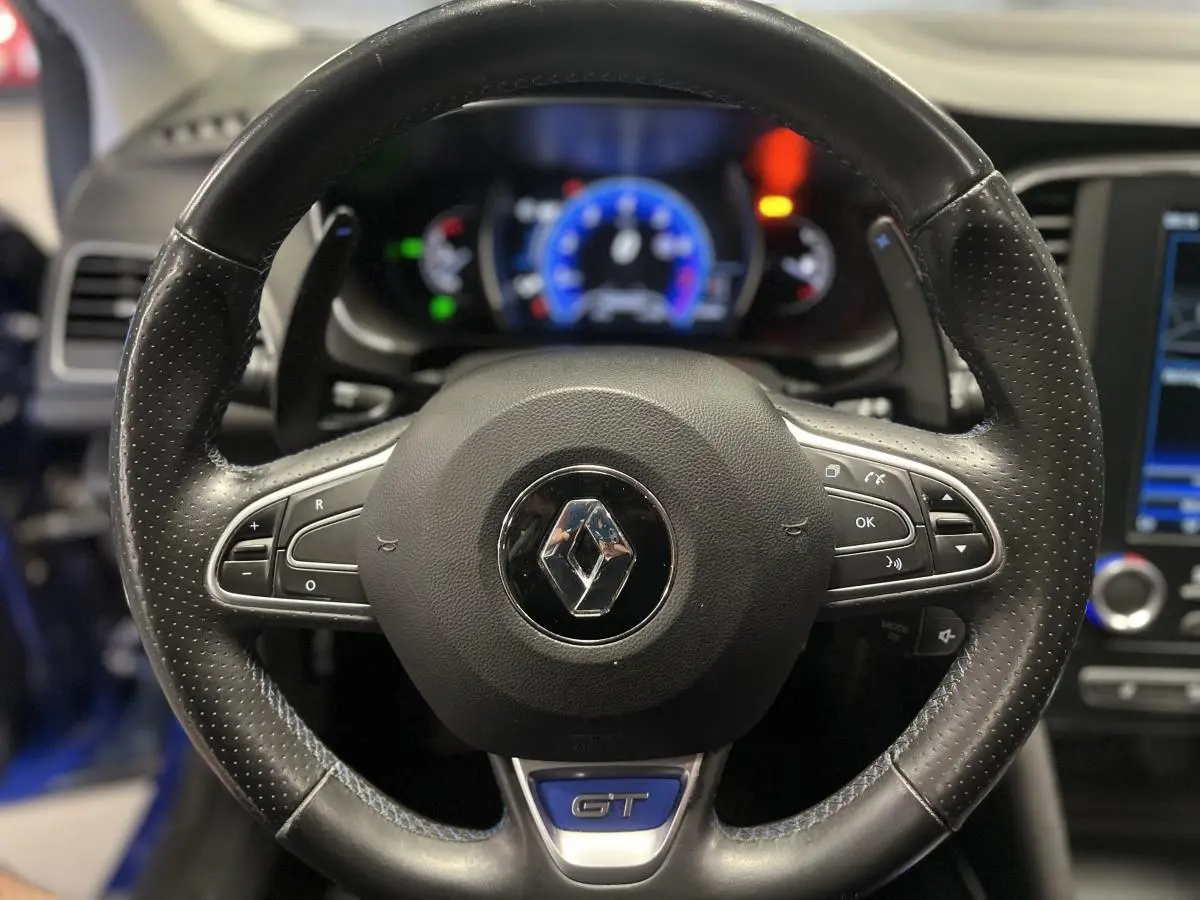 Gros plan sur le volant cuir perforé noir avec logo Renault et badge GT, tableau de bord numérique en arrière-plan.