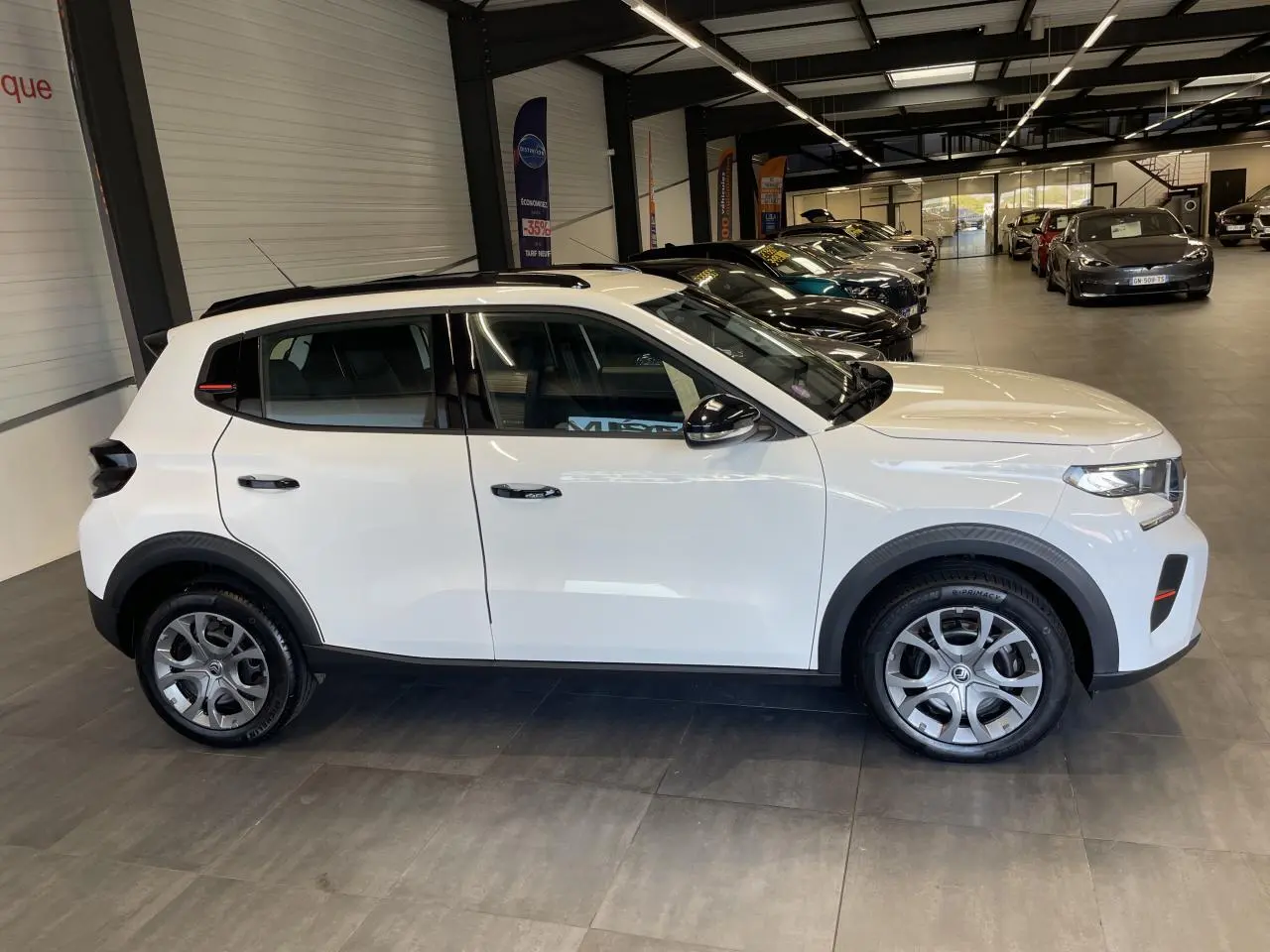 Vue de profil côté gauche d'une Citroën C3 blanche avec barres de toit noir brillant en showroom.