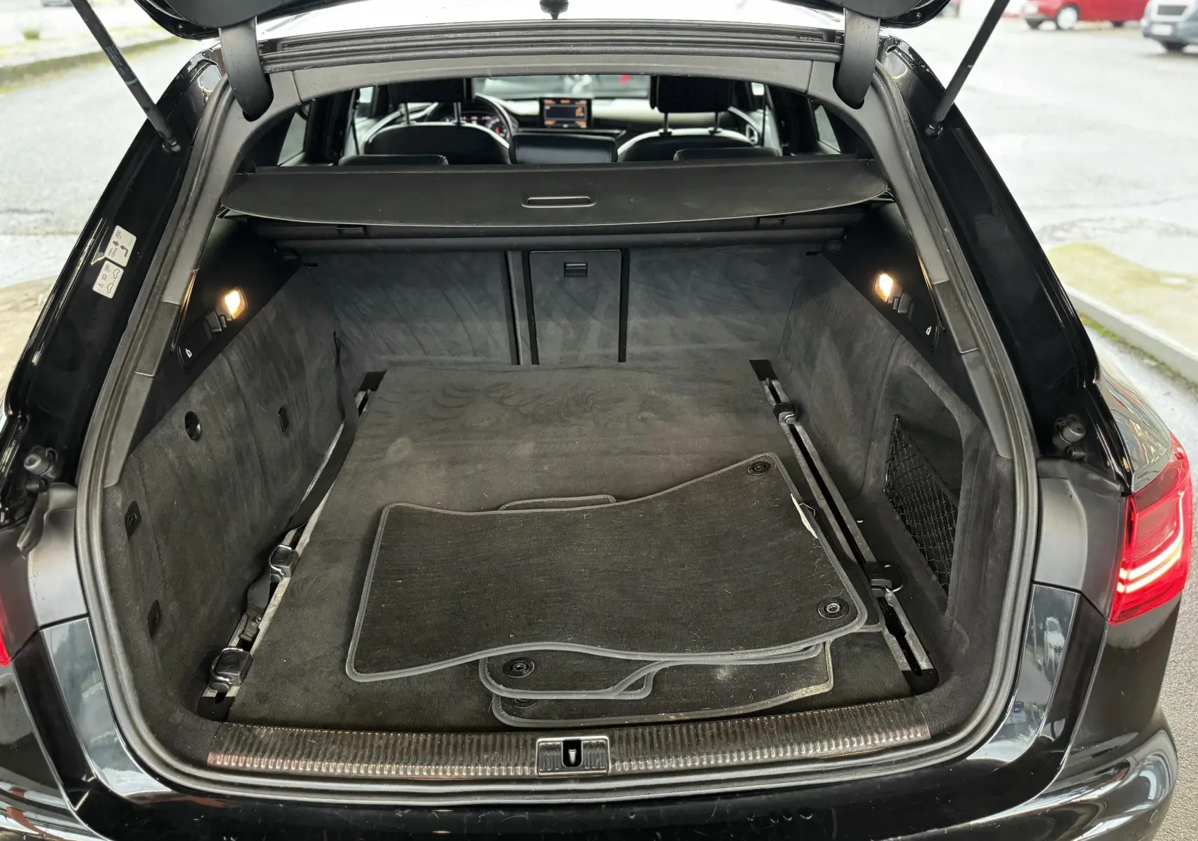 Vue arrière du coffre ouvert d'une Audi A6 Avant Allroad noire, avec tapis de sol noirs et intérieur spacieux.