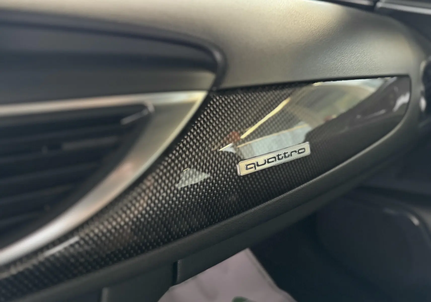 Détail intérieur côté passager montrant la finition carbone brillante avec le logo Quattro sur tableau de bord noir.