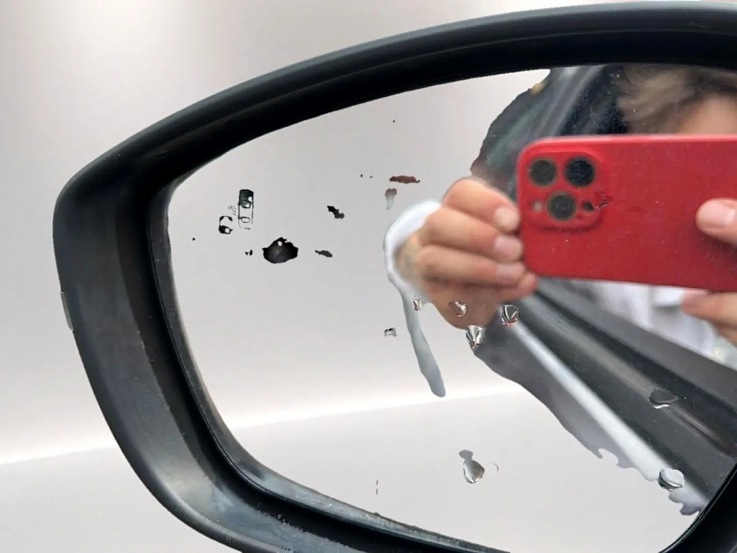 Rétroviseur extérieur gauche d’un Opel Mokka gris 2025 avec traces d’eau et reflet d’une main tenant un smartphone rouge.