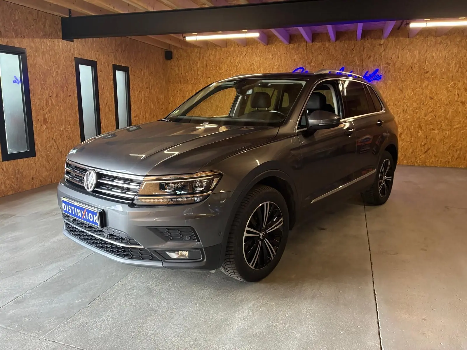 Volkswagen Tiguan gris 3/4 avant droit, avec jantes noires et détails chromés, en intérieur sur sol béton.