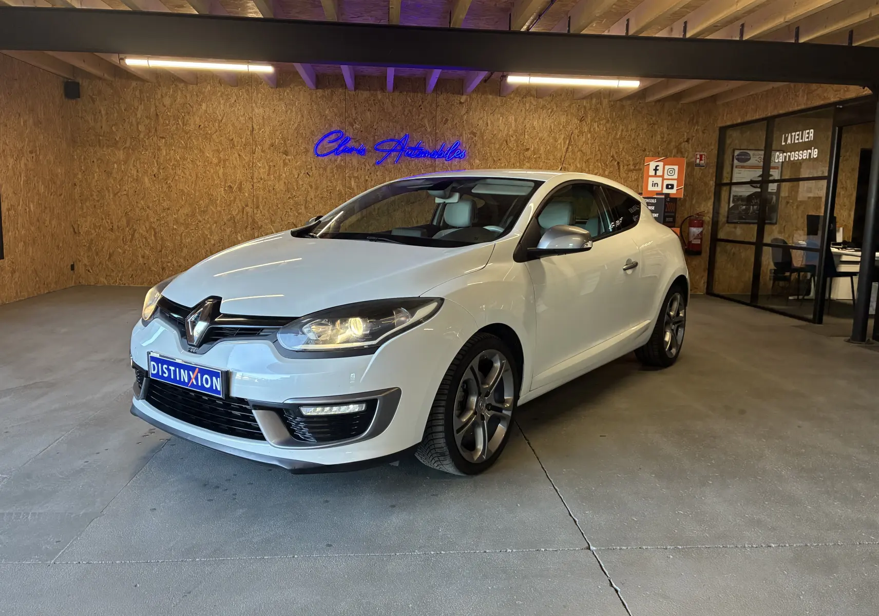 Renault Mégane Coupé blanc vue 3/4 avant droit en intérieur avec phares allumés et jantes alliage.