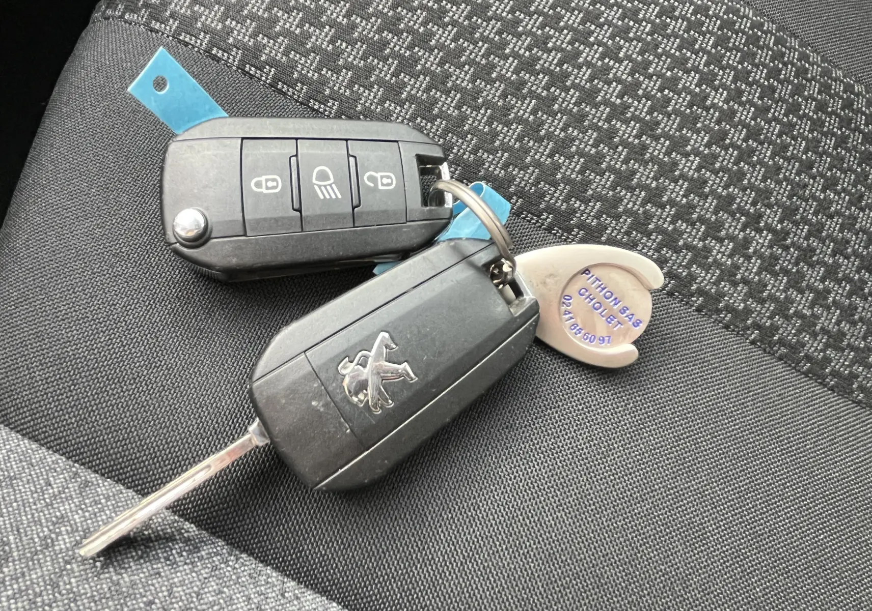 Clé de voiture Peugeot noire posée sur un siège tissu gris foncé à motifs, avec porte-clés métallique et télécommande.