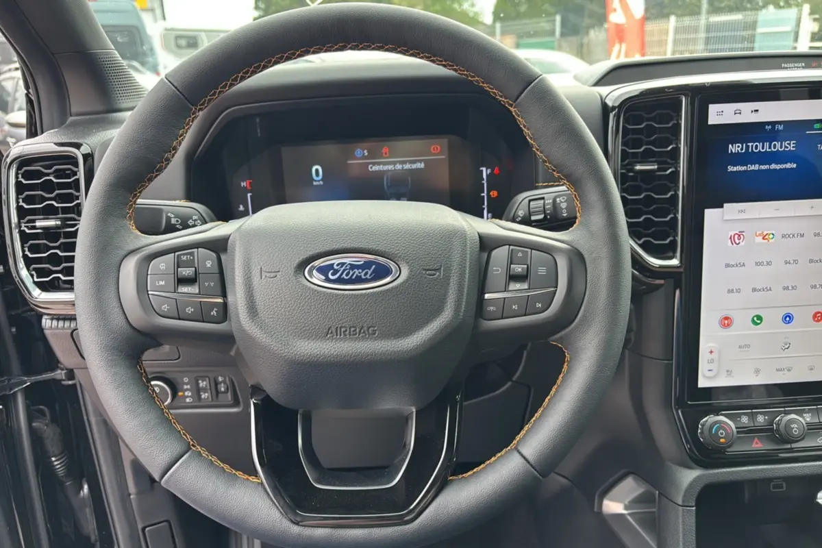 Vue rapprochée du volant cuir noir avec surpiqûres orange du Ford Ranger 2025, tableau de bord et écran tactile visibles.