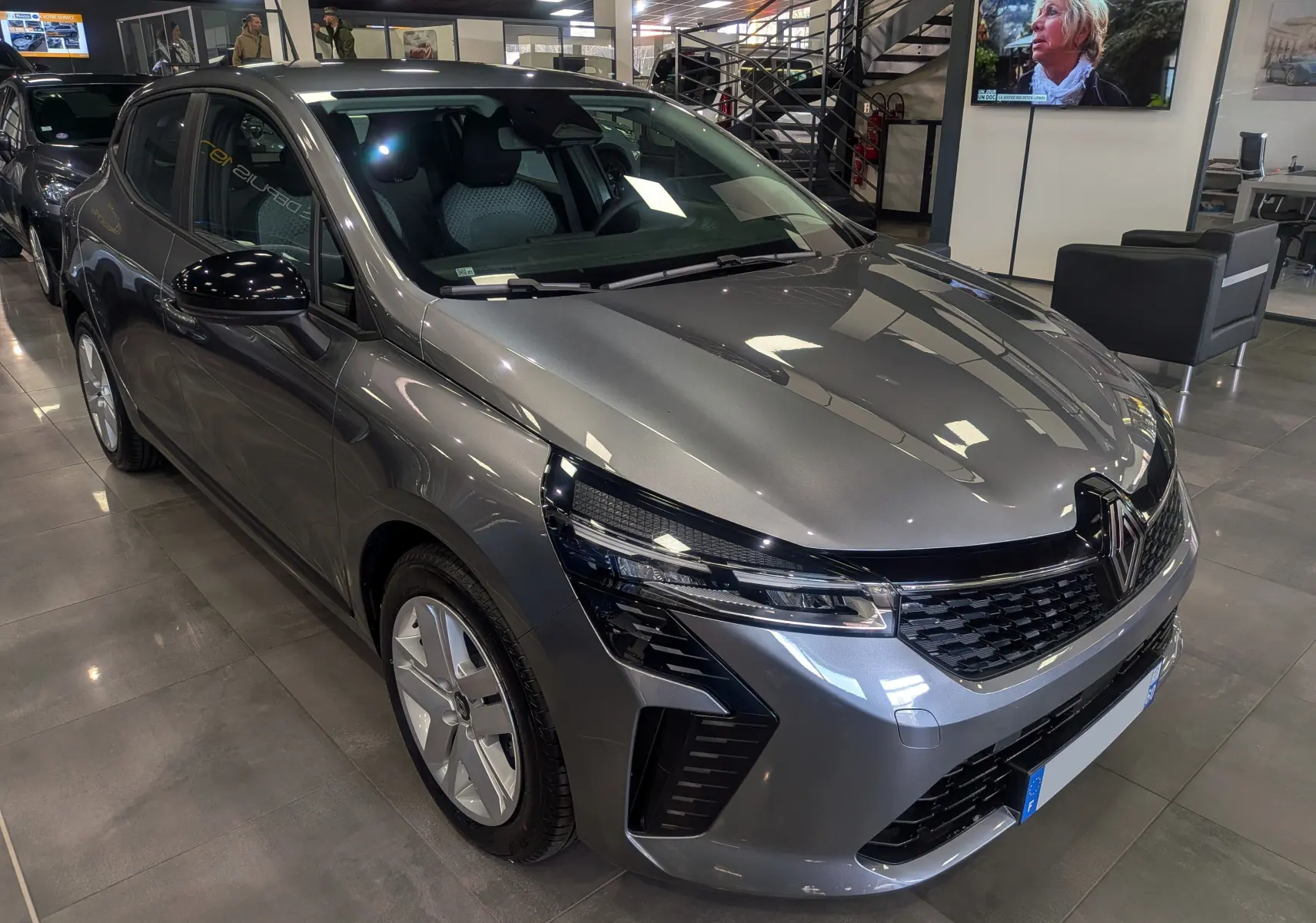Renault Clio V Blue dCi 100 Evolution gris schiste vue 3/4 avant droit en intérieur showroom, calandre et phares modernes.