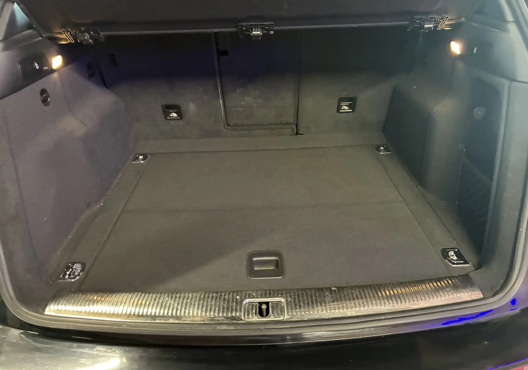 Coffre ouvert vu de l'arrière d'un Audi SQ5 noir, avec tapis de sol et éclairage intérieur latéral allumé.