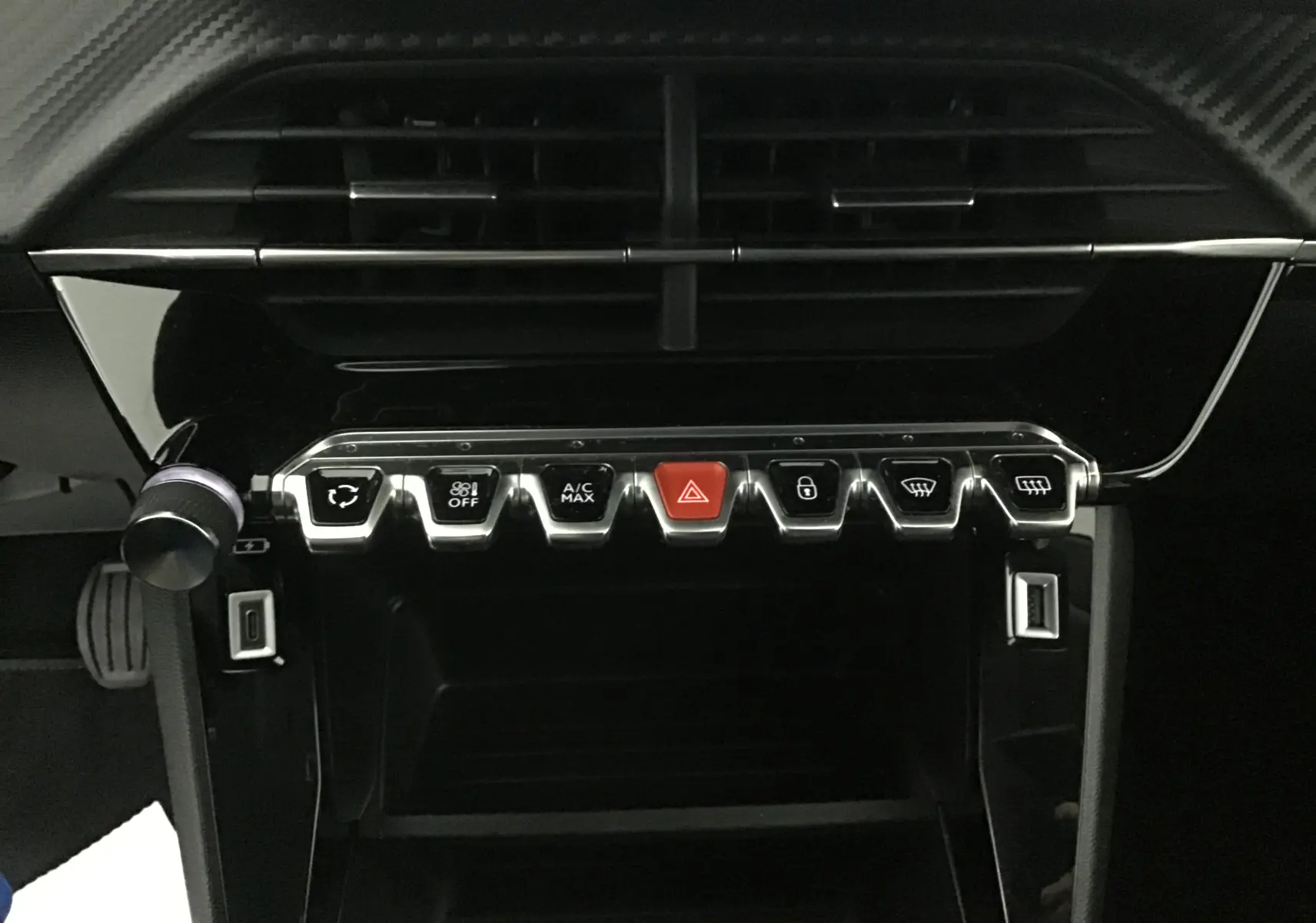 Gros plan sur la console centrale noire brillante du Peugeot 208 2022 avec boutons chromés et bouton rouge d'alerte.