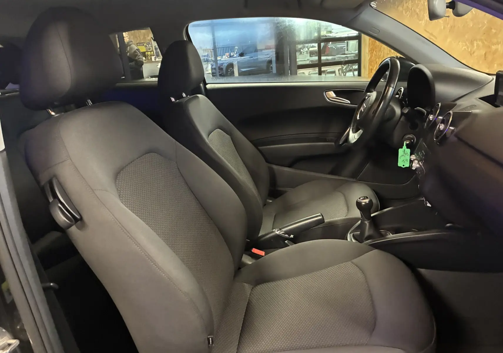 Vue intérieure côté gauche de l'Audi A1 noire de 2016, montrant les sièges en tissu gris-noir et le volant avec boîte manuelle.