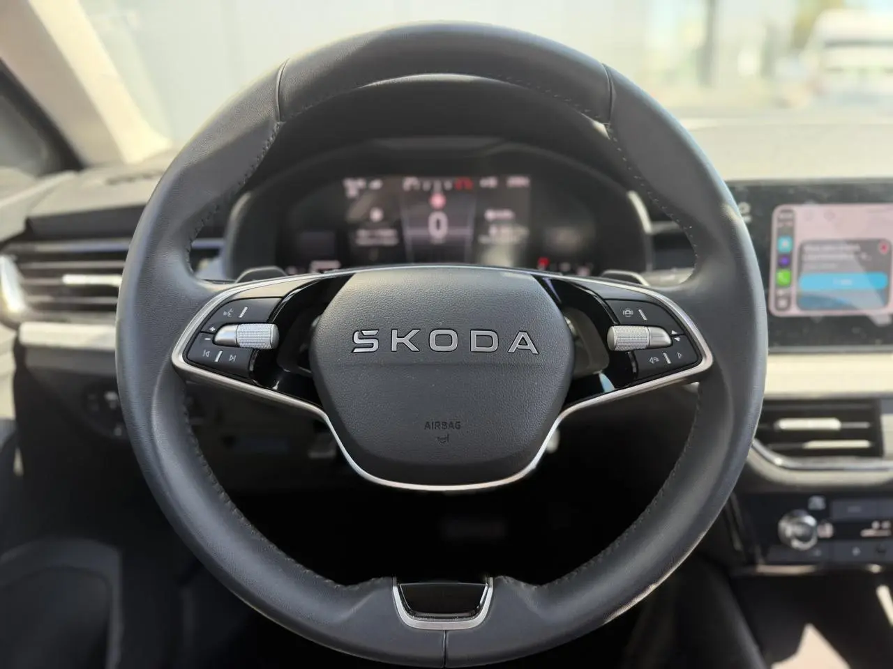 Volant cuir multifonctions SKODA en vue frontale avec tableau de bord numérique et écran tactile à droite flou.