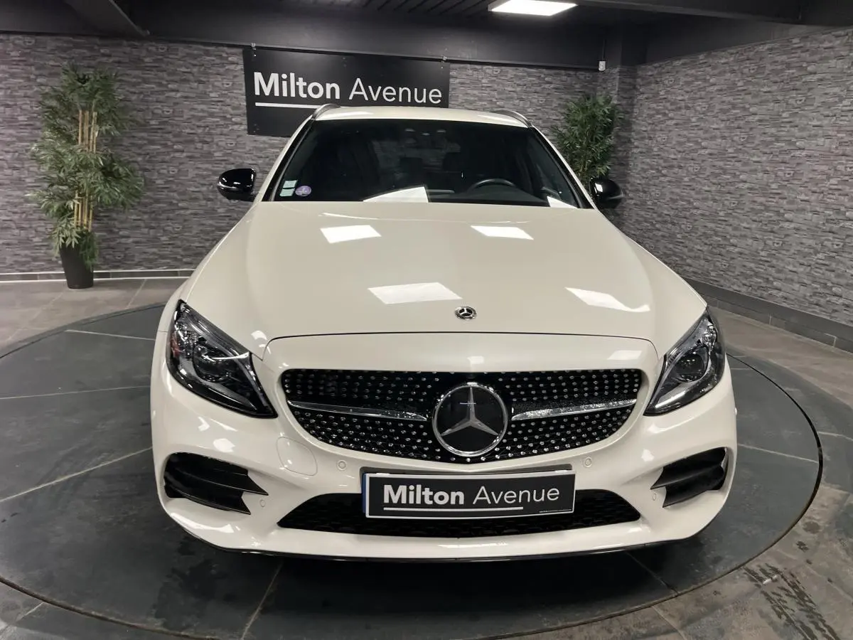 Vue frontale d'un break Mercedes Classe C blanc avec calandre AMG et logo étoile au centre, en intérieur showroom.