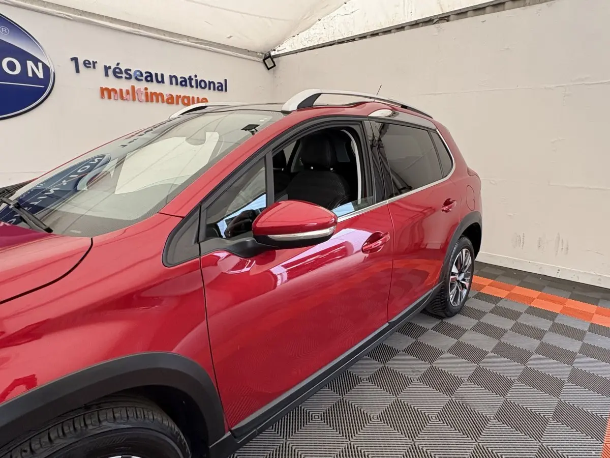 Vue 3/4 avant droit d'un Peugeot 2008 rouge brillant avec rétroviseurs chromés et barres de toit chromées.