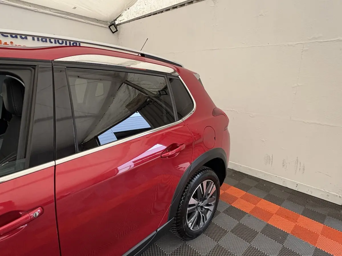 Vue côté droit arrière du Peugeot 2008 rouge avec jantes alliage et barres de toit chromées dans un garage.