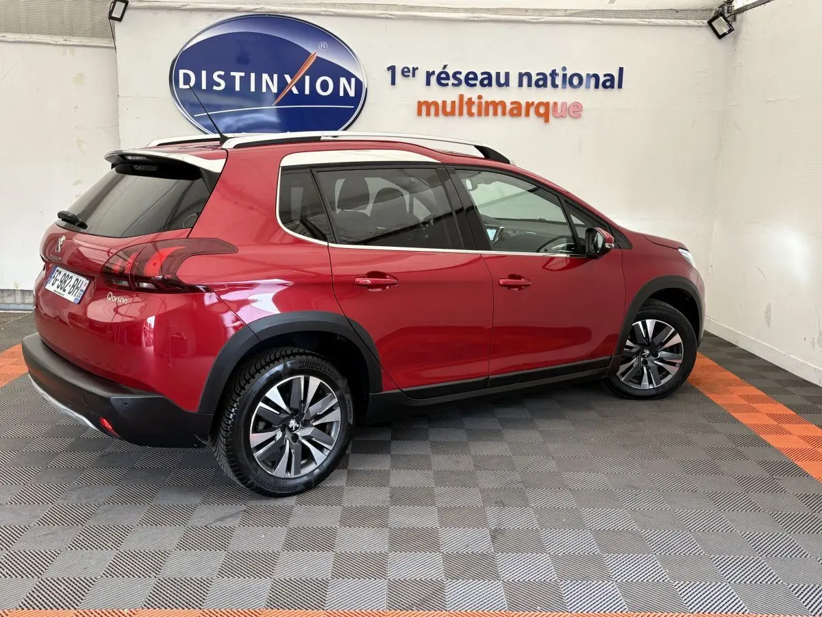 Vue 3/4 arrière droite d'un Peugeot 2008 rouge 2019 avec jantes alliage et barres de toit chromées dans un showroom.