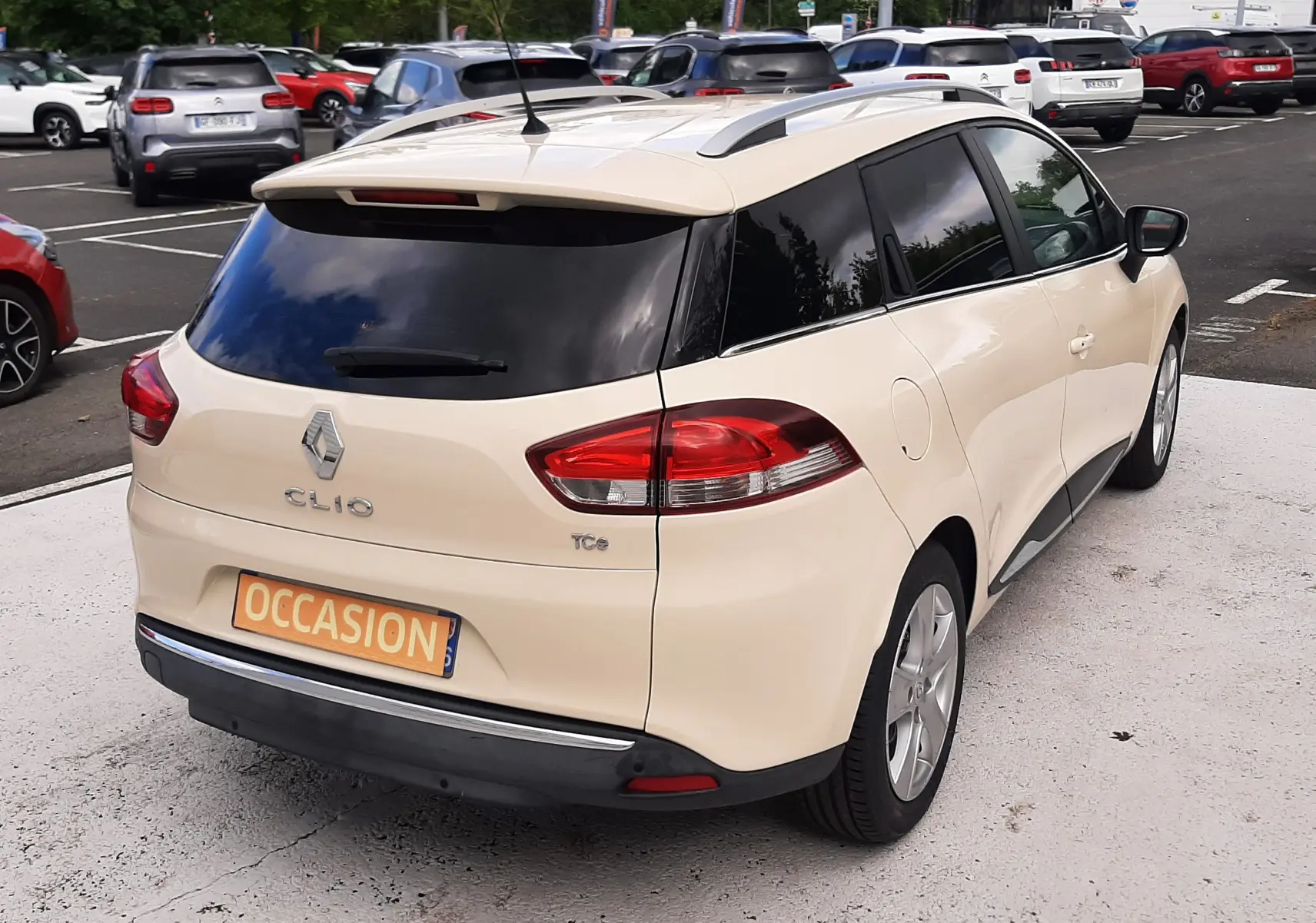 Vue 3/4 arrière droite d'une Renault Clio IV Estate beige avec vitres teintées et toit avec barres de toit.