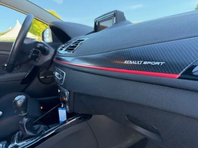 Intérieur côté passager de la Renault Mégane III Coupé RS blanc nacré, tableau de bord carbone avec bande rouge et logo Renault Sport.