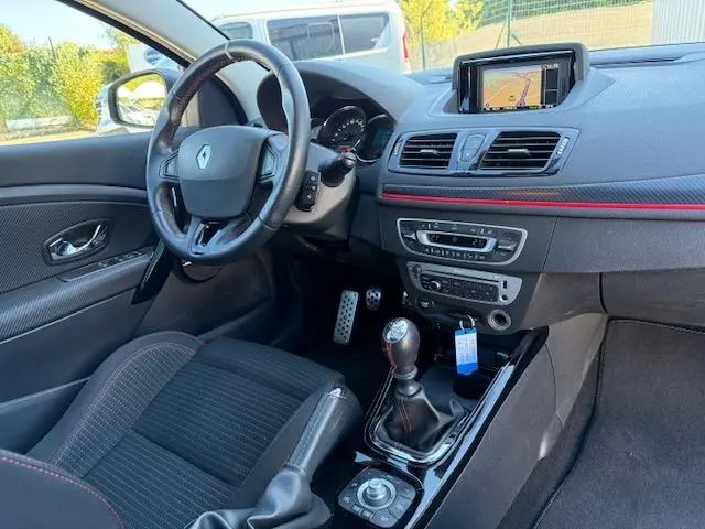 Intérieur de Renault Mégane III Coupé 2.0T blanc nacré, vue côté conducteur, volant cuir, levier de vitesse manuel et écran GPS intégré.