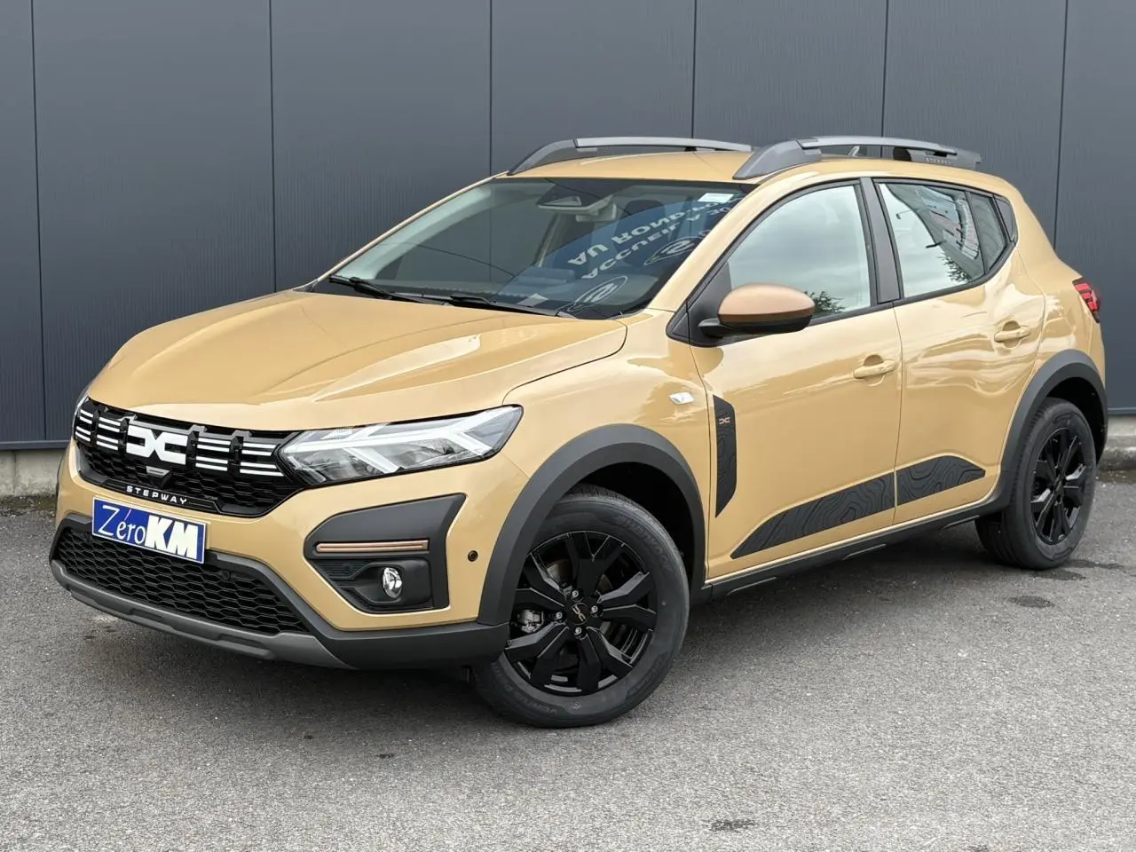 Vue 3/4 avant droite d'une Dacia Sandero Stepway beige Safari avec jantes noires et barres de toit gris Mégalithe.