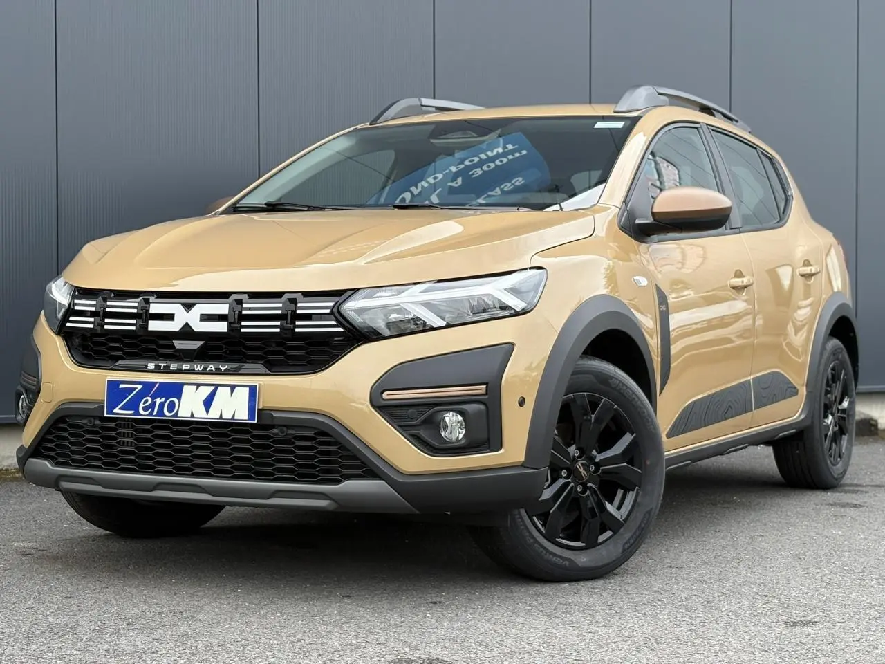 Vue 3/4 avant droit d'une Dacia Sandero Stepway 2025 beige Safari avec jantes noires et barres de toit gris Mégalithe.