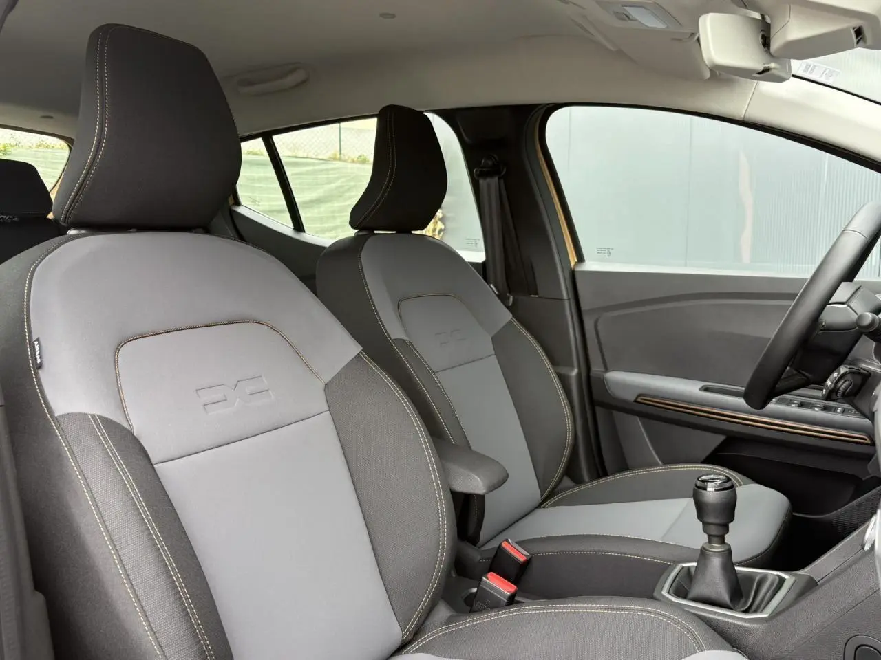 Vue intérieure côté droit des sièges avant gris clair et foncé avec accoudoir, volant et levier de vitesses manuelle visibles.