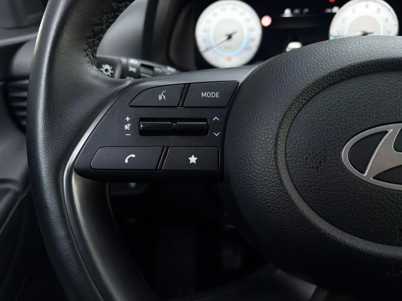 Gros plan sur les commandes gauche du volant cuir noir de la Hyundai i20 2025 avec compteurs numériques en arrière-plan flou.