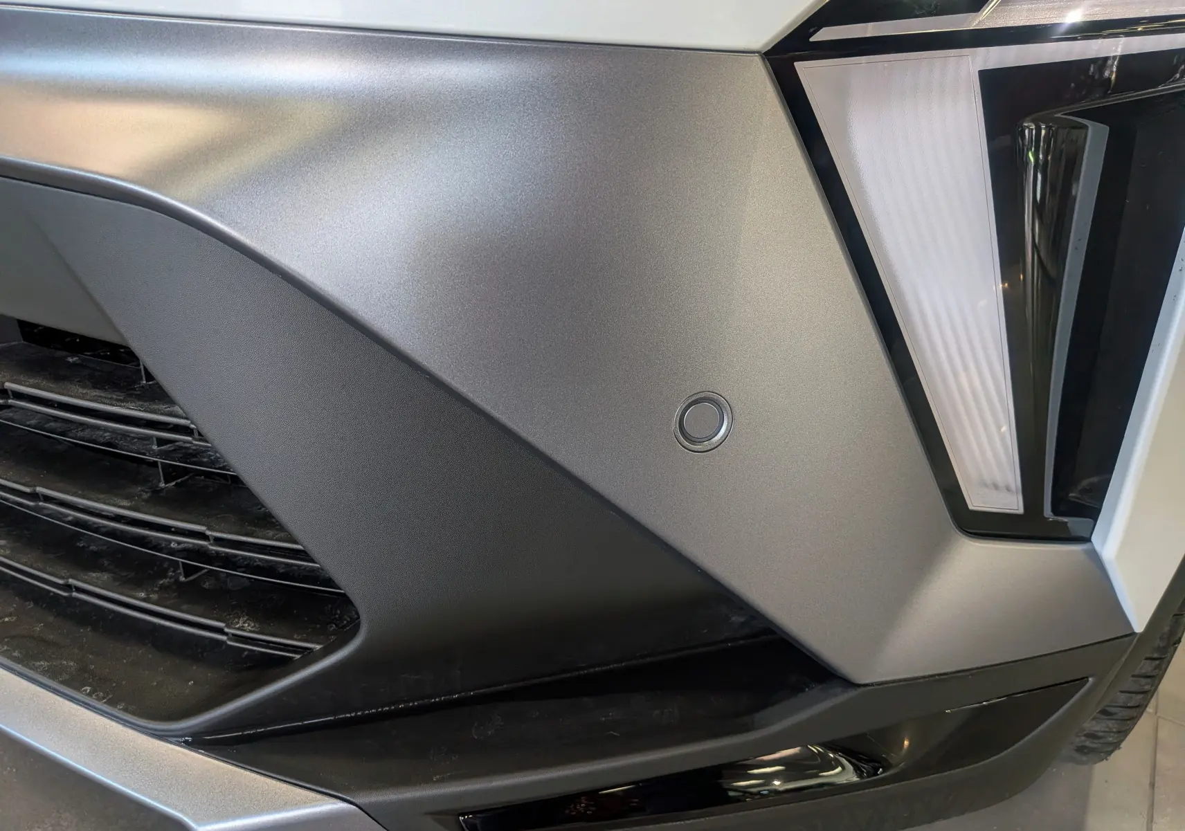 Gros plan sur le pare-chocs avant droit gris mat du Renault Symbioz blanc nacré, avec capteur de stationnement circulaire visible.