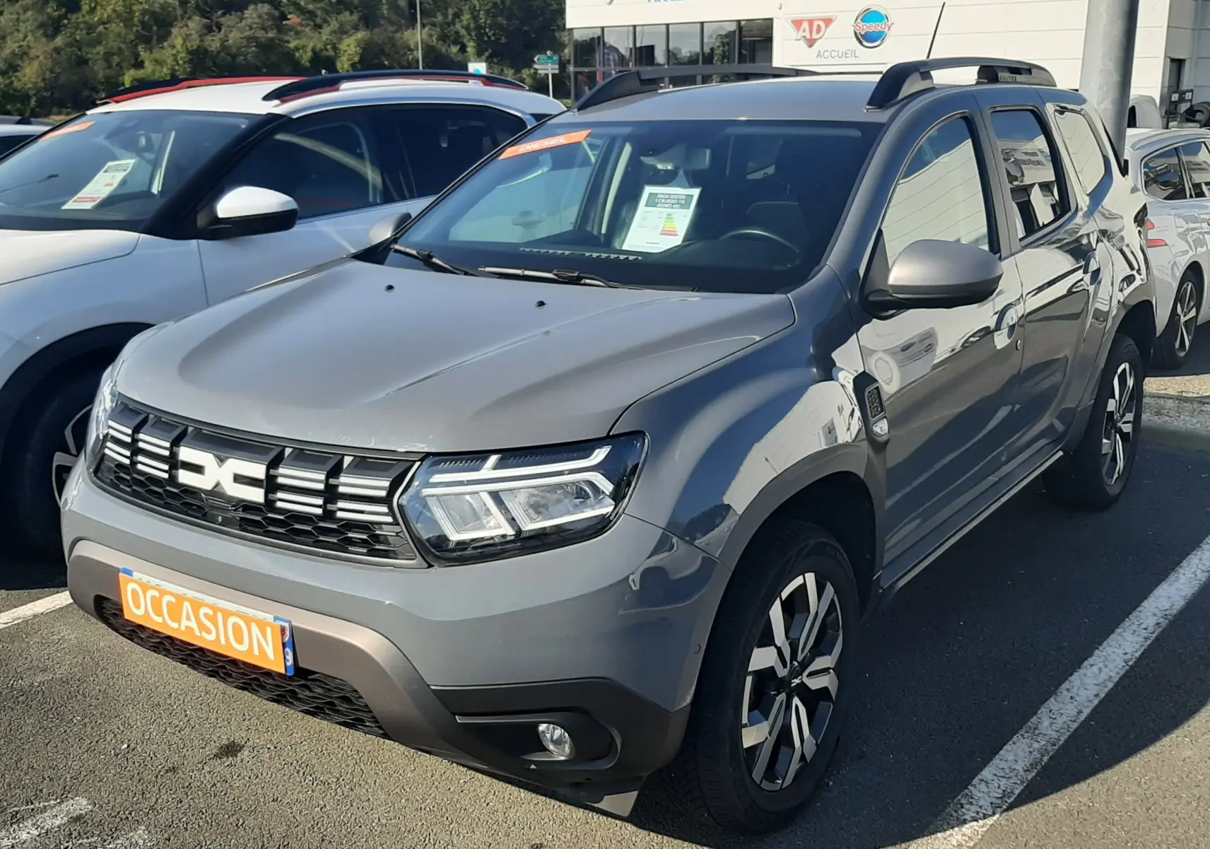 Dacia Duster gris 2024 en 3/4 avant droit, avec jantes bi-ton et plaques "Occasion" visible au parking.