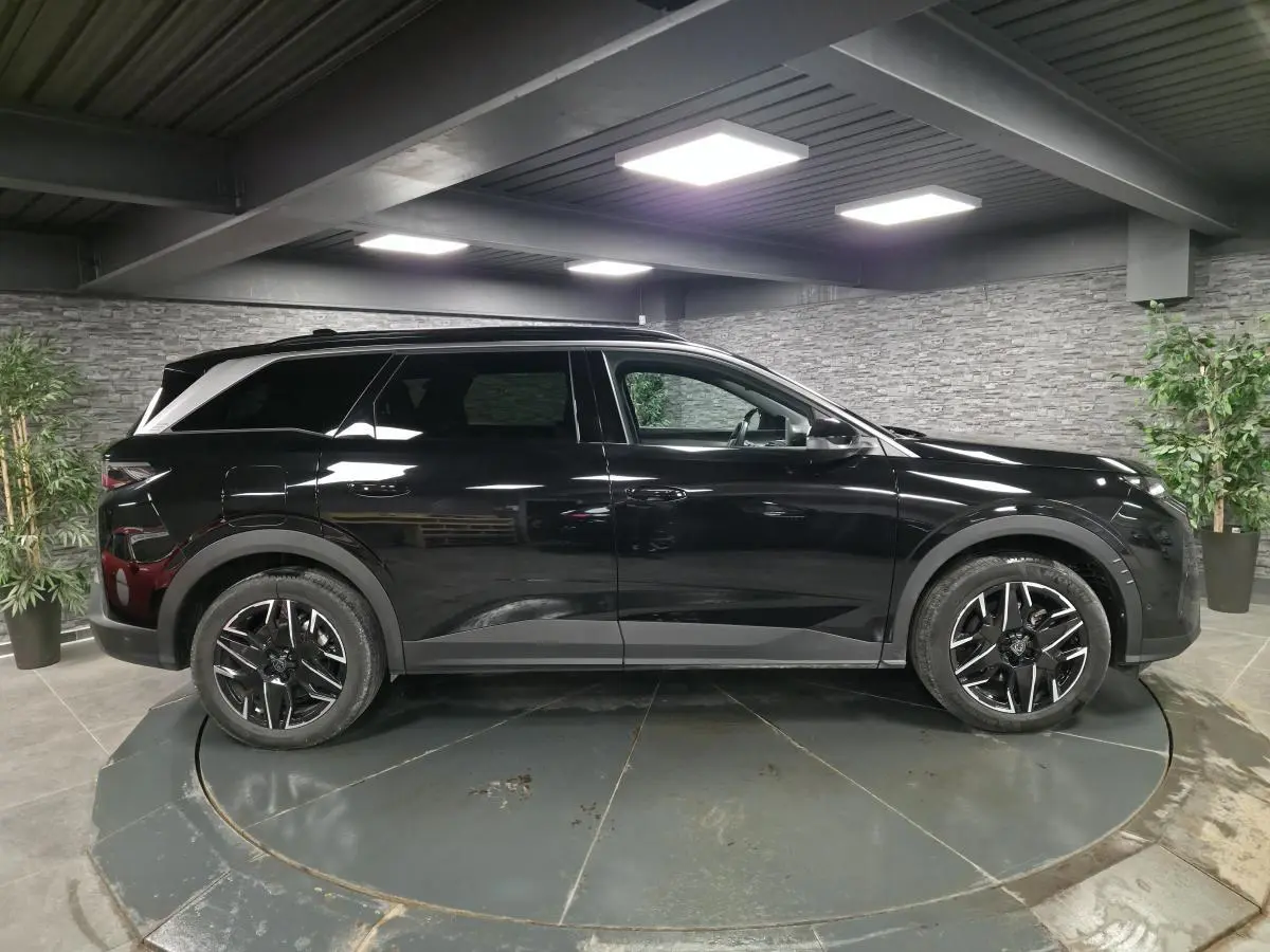 Vue de profil côté gauche d’un Peugeot 5008 noir 2025 avec jantes alliage noires et barres de toit noires brillantes dans un showroom.