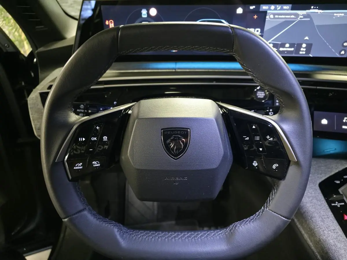 Vue rapprochée du volant compact cuir noir avec commandes intégrées et logo Peugeot sur fond intérieur moderne.