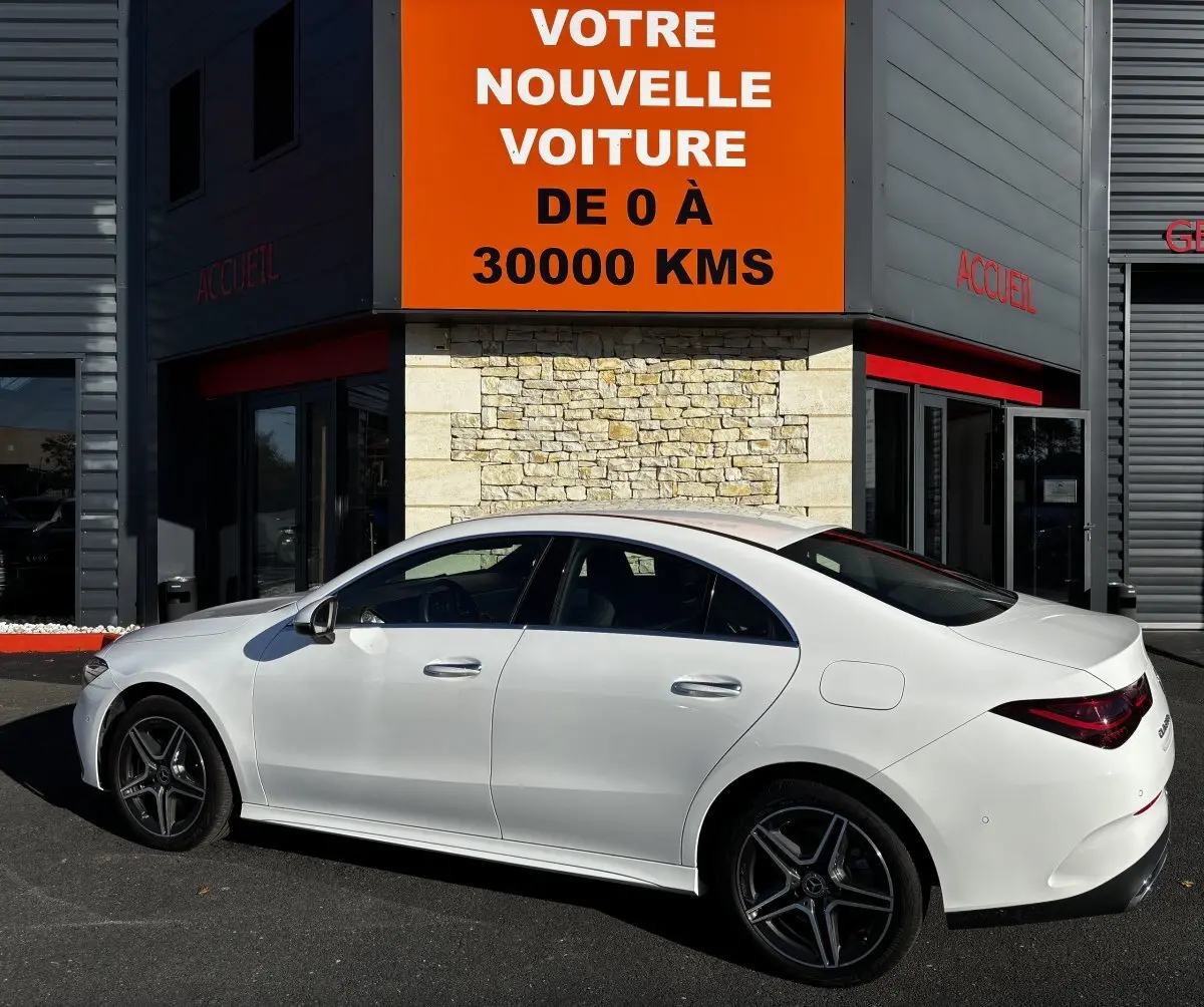 Vue de profil côté gauche d'une Mercedes CLA 250 e AMG Line blanche stationnée devant un bâtiment avec panneau orange.