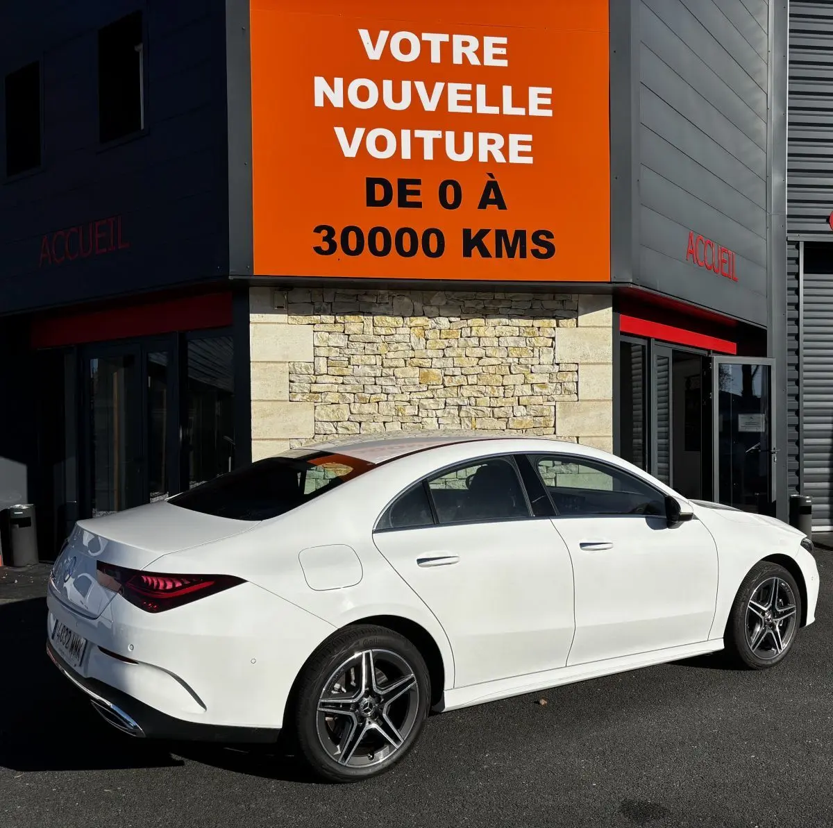 Vue 3/4 arrière droite d'une Mercedes CLA 250 e AMG Line blanche stationnée devant un bâtiment avec panneau orange.