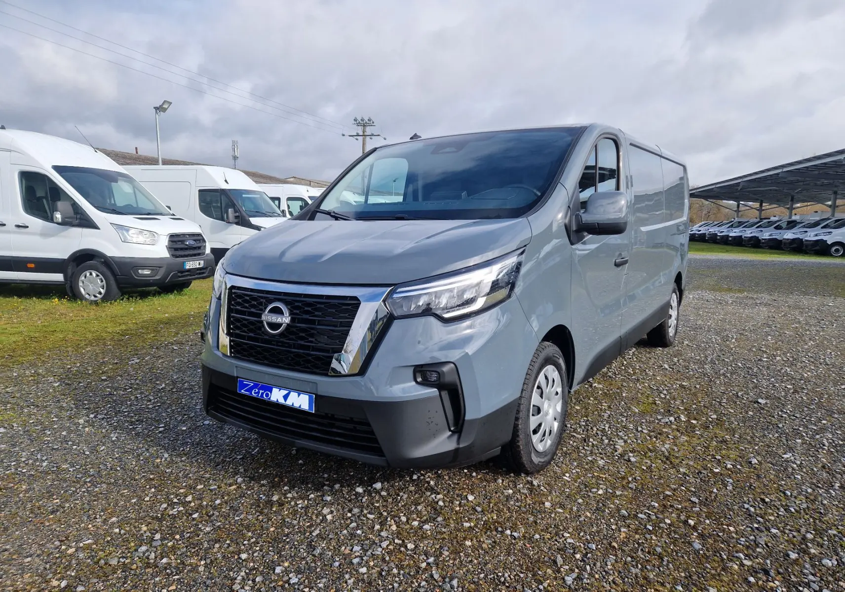 Nissan Primastar gris urbain vue 3/4 avant droit sur parking avec plusieurs fourgons blancs en arrière-plan.