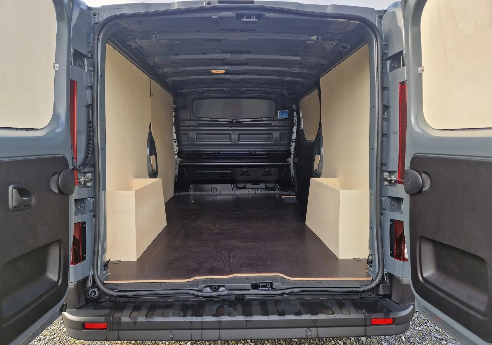 Vue arrière ouverte du Nissan Primastar gris urbain 2025, montrant l'espace de chargement avec aménagements bois clairs.