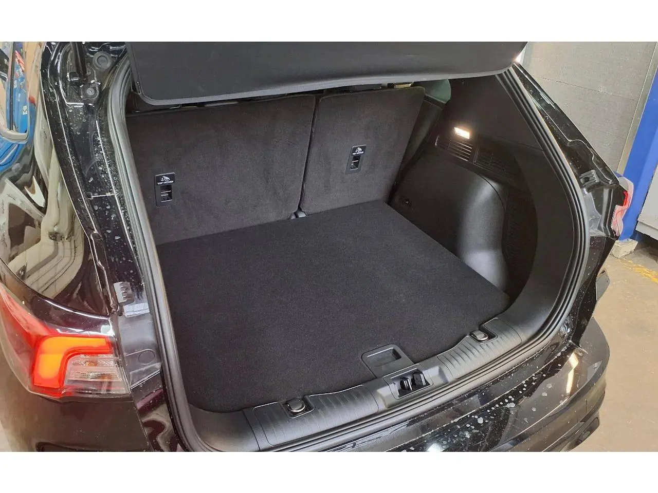 Coffre ouvert vu de l'arrière droit du Ford Kuga noir Agathe 2024 avec plancher de coffre plat et éclairage intérieur.