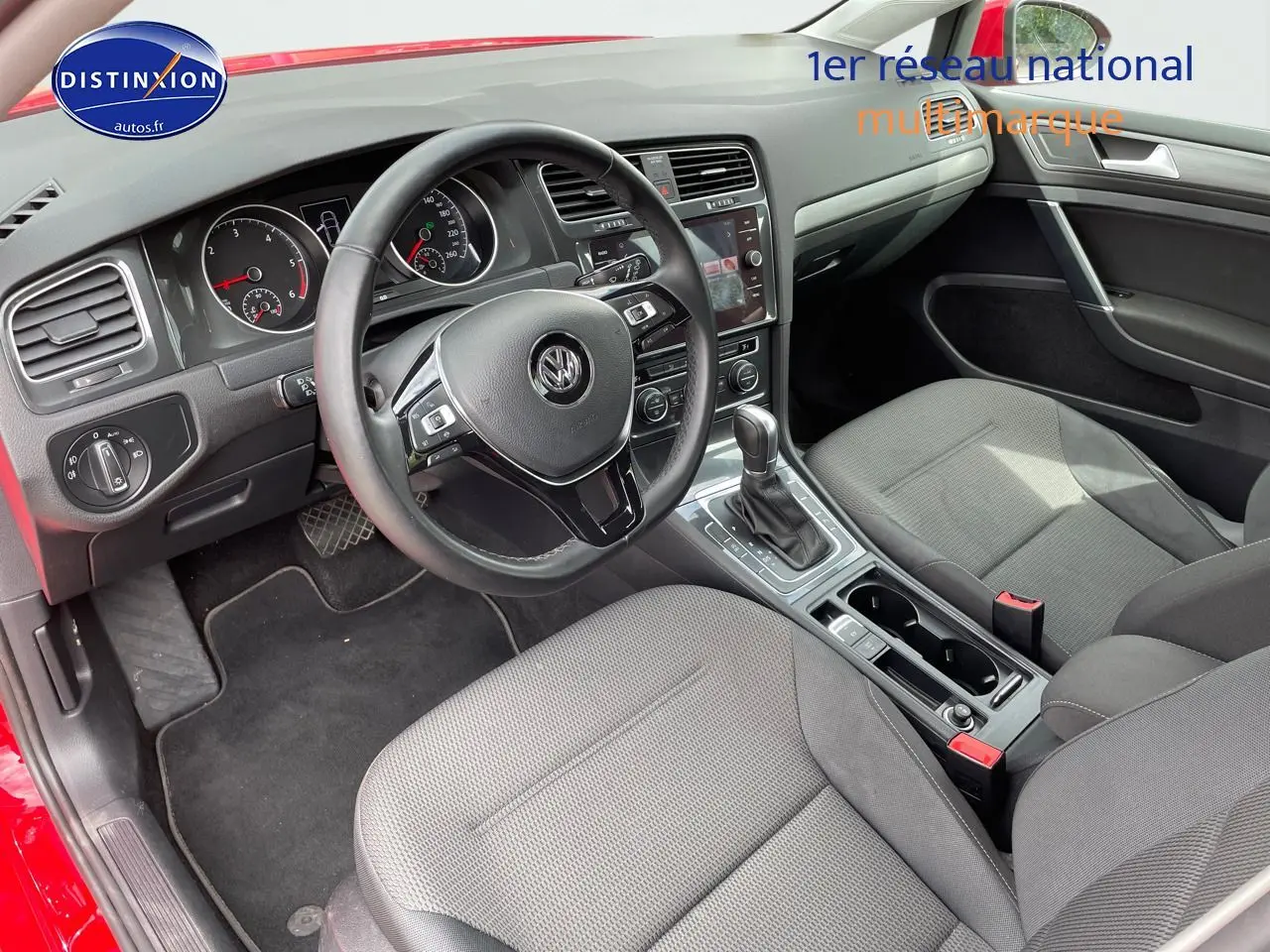 Vue intérieure côté conducteur d'une Volkswagen Golf rouge, montrant le volant, le tableau de bord et les sièges en tissu gris.