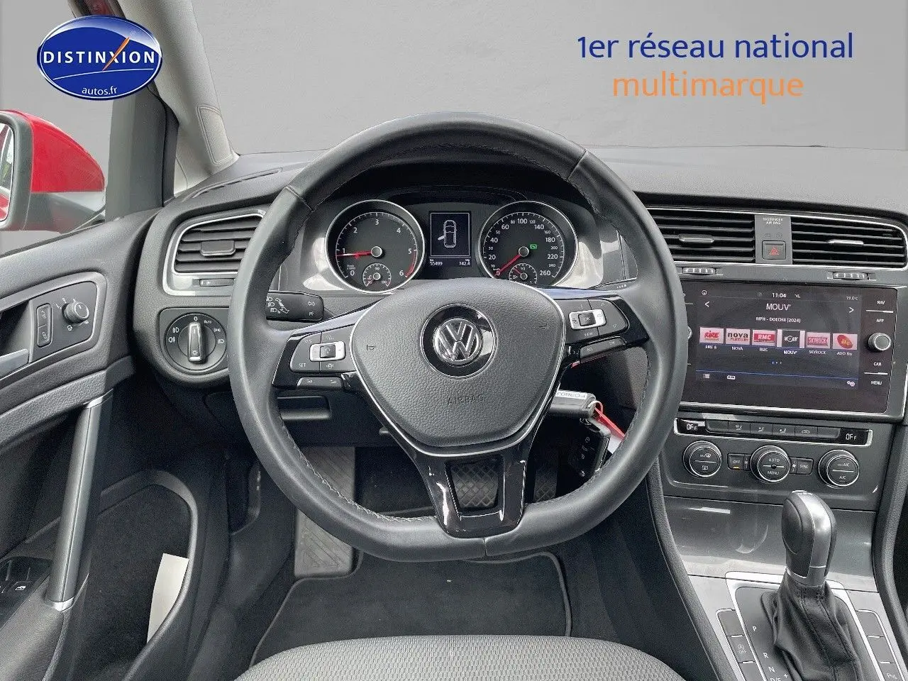 Vue intérieure centrée sur le volant et le tableau de bord d’une Volkswagen Golf rouge, avec écran tactile et commandes visibles.
