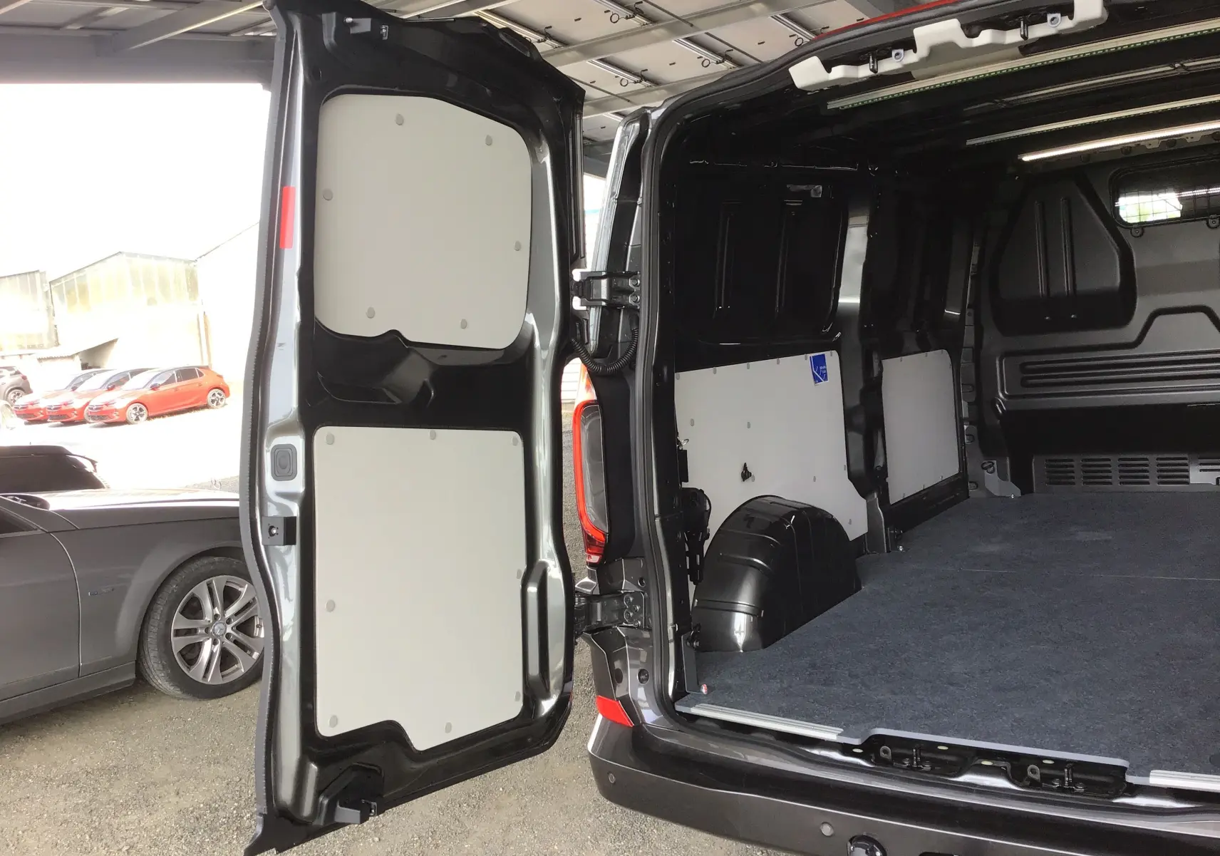 Vue arrière ouverte du Volkswagen Transporter T7 gris anthracite montrant l'espace de chargement vide avec habillage intérieur noir et panneaux blancs.