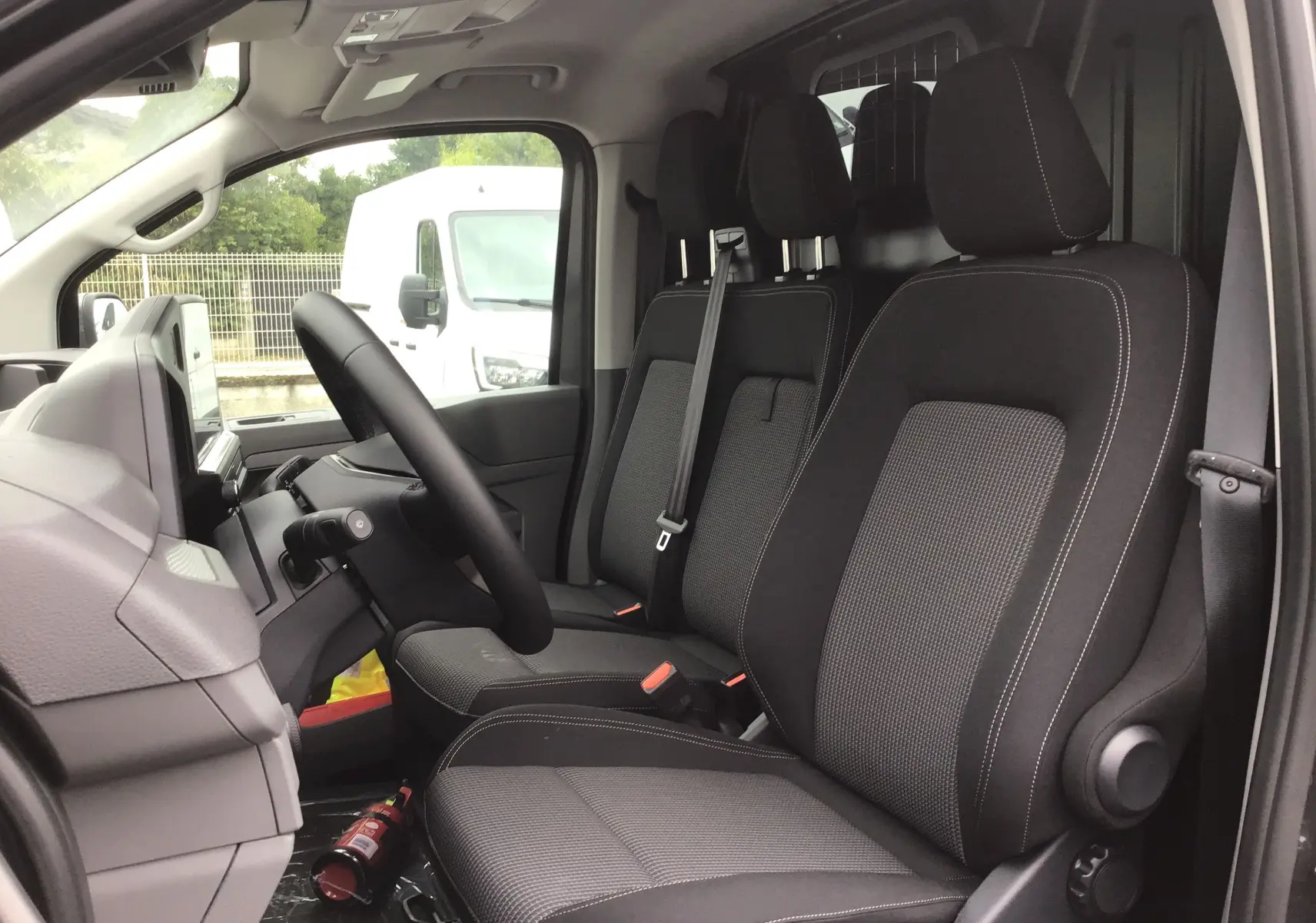 Vue intérieure côté conducteur du Volkswagen Transporter T7 2025, sièges tissu gris anthracite avec appuie-têtes et ceintures visibles.