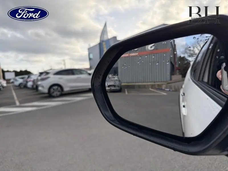 Rétroviseur côté droit d'une Toyota Yaris Cross blanc lunaire nacré avec gouttes d'eau, vue sur parking extérieur.