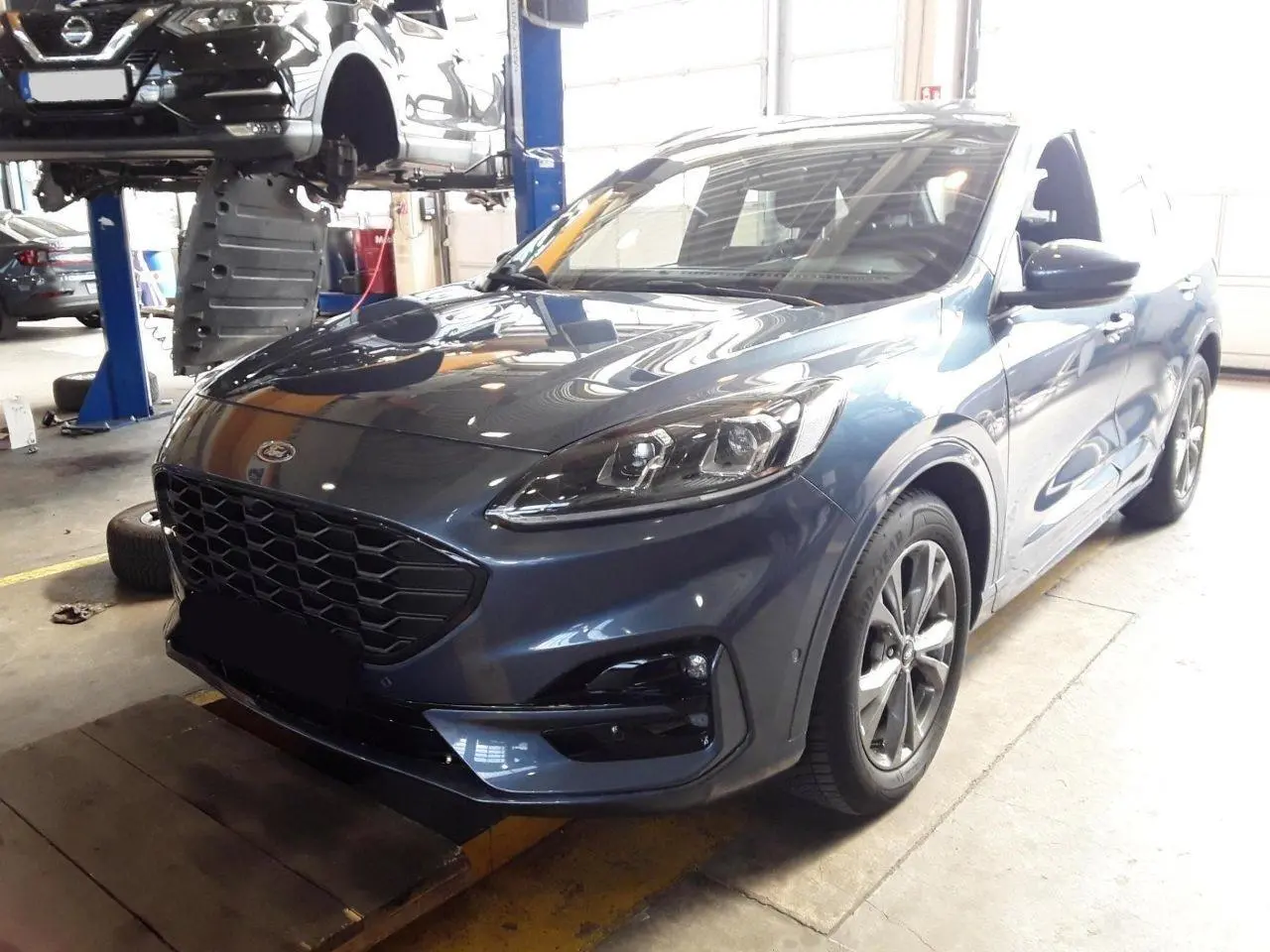 Ford Kuga 2024 bleu azur en 3/4 avant droit, avec calandre noire et jantes alliage visibles en atelier.