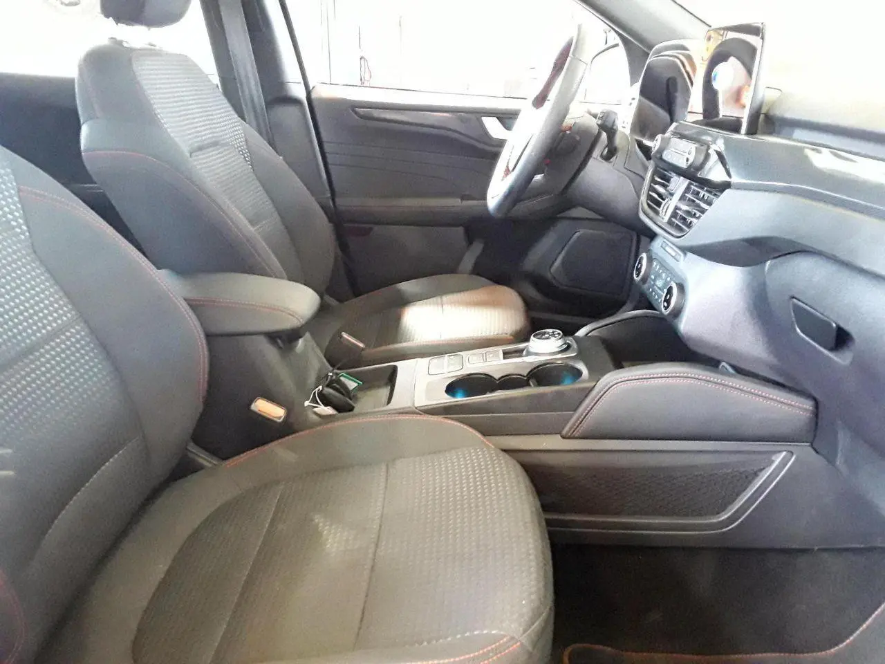 Vue intérieure côté conducteur du Ford Kuga 2024, sièges gris avec surpiqûres rouges et console centrale moderne.